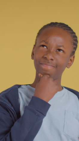 Vertical-Video-Of-Young-Boy-Thinking-Against-Yellow-Background-In-Studio